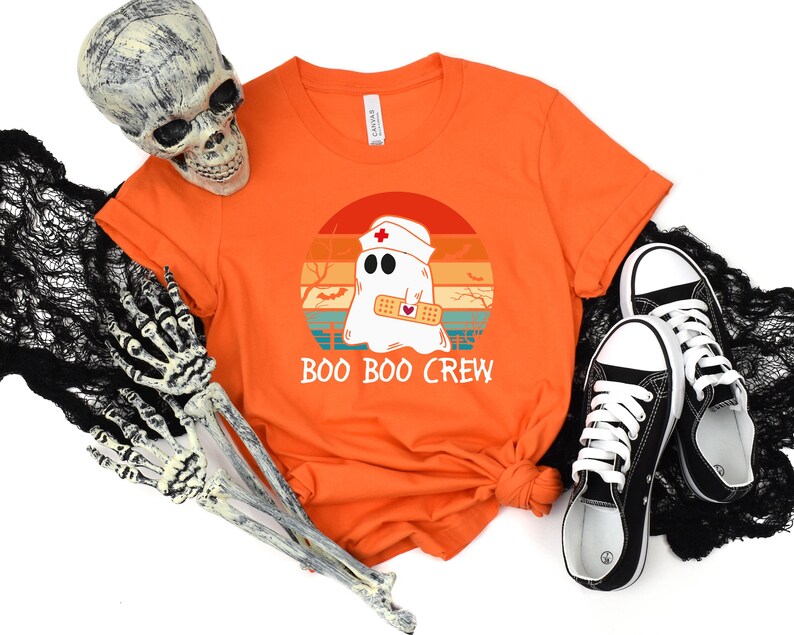 May include: Orange t-shirt with a graphic of a ghost wearing a nurse's cap and a bandage, set against a sunset design. The text "BOO BOO CREW" is below the graphic. Black and white sneakers are next to the shirt.