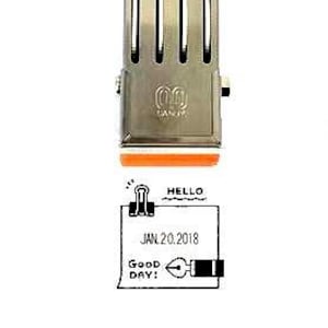 May include: A silver-toned metal date stamp featuring a rectangular body with four vertical slots and an orange base. The stamp imprint displays a binder clip icon, the word HELLO, the date JAN. 20. 2018, the text GOOD DAY, and a fountain pen nib icon.