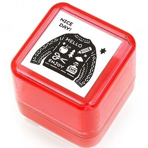May include: A square red rubber stamp featuring a white top label with a black graphic of a sweater. The sweater design includes the text NICE DAY, HELLO, and ENJOY INK, along with small icons of an apple, a pencil, scissors, a cloud, and a star.