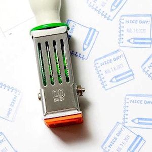May include: A metal self-inking stamp with a white handle, green ring, and orange base. The stamp features the brand name SANY and creates a blue ink impression of a spiral-bound notebook with a pencil and the text NICE DAY! JUL 16 2021.