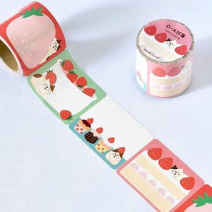 May include: Decorative washi tape featuring a repeating pattern of strawberries, cakes, and a cat. The tape is pink, red, green, and white. One roll is unrolled, showing the pattern, and another roll is intact.