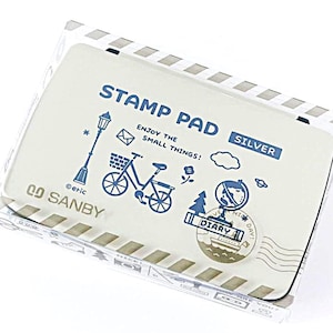 May include: A rectangular silver stamp pad in a clear plastic case with a white lid. The lid features blue illustrations of a street lamp, a bicycle, a globe, and the text STAMP PAD, SILVER, SANBY, and ENJOY THE SMALL THINGS! with a DIARY stamp.
