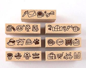 May include: A set of seven rectangular wooden rubber stamps featuring dark brown line art illustrations of daily life objects, including a pen, envelope, coffee cup, glasses, books, a dog, a cafe menu, flowers, a shopping bag, and a car.