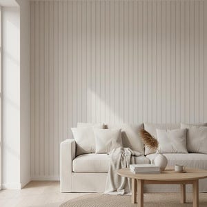 May include: A neutral-toned living room scene featuring a beige sofa with several white pillows and a matching throw. A round wooden coffee table holds books, a vase, and a mug. The room has a light wood floor, a woven rug, and a vertical-striped wall.