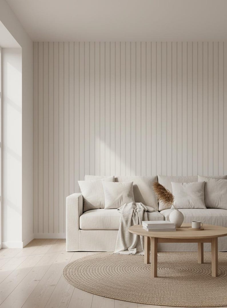 May include: A neutral-toned living room scene featuring a beige sofa with several white pillows and a matching throw. A round wooden coffee table holds books, a vase, and a mug. The room has a light wood floor, a woven rug, and a vertical-striped wall.