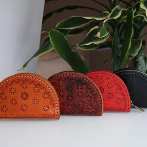 Puede incluir: Una colección de cuatro monederos de cuero en varios colores: naranja, marrón, rojo y negro. Cada monedero tiene forma de semicírculo, cierre de cremallera y un patrón decorativo de estrellas. Los monederos están alineados.