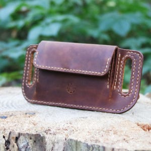 Peut inclure: Une pochette de ceinture en cuir marron avec fermeture à rabat et coutures décoratives. La pochette présente un petit motif en relief et des passants de ceinture sur les côtés. L'article est posé sur une surface en bois.
