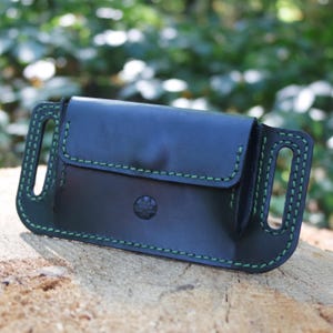 Puede incluir: Bolsa de cinturón de cuero negro con costuras verdes. La bolsa tiene un cierre de solapa y un botón decorativo. El diseño incluye presillas para cinturón para una fácil fijación. El artículo se muestra sobre una superficie de madera.