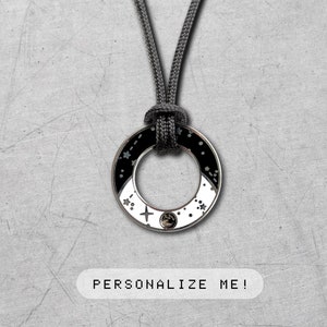 Puede incluir: Un collar negro y blanco con un colgante redondo que presenta un círculo blanco con puntos negros y un pequeño diamante en el centro. El colgante está unido a un cordón negro. El texto "¡PERSONALÍZAME!" está impreso debajo del colgante.