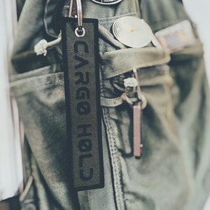 May include: Close-up of a green military-style backpack with multiple pockets and zippers. A black tag with the text "CARGO HOLD" hangs from a silver keyring. The backpack has several patches, including a circular one with the text "RESISTANCE IS FUTILE".