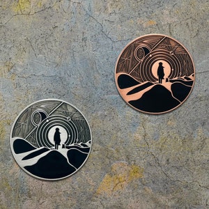 May include: Two circular metal pins featuring a black and white design of a person standing in a desert landscape with a starry sky and planets in the background. One pin is silver and the other is copper.