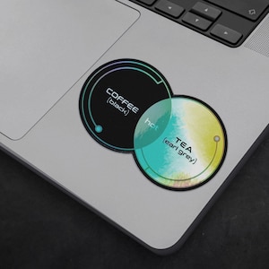 Puede incluir: Una pegatina en blanco y negro con el texto "COFFEE (black)" y una pegatina azul verdoso y amarilla con el texto "TEA (earl grey)". Las pegatinas se superponen y el texto "hot" está escrito en el área superpuesta.