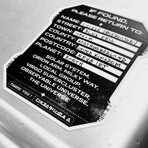 May include: A black and white sticker with the text "If Found, Please Return To: Kielder Observatory, Black Fell, Hexham, Northumberland, United Kingdom, NE48 1EJ, Planet: Earth." The sticker also lists the Solar System, Orion Arm, Milky Way, Virgo Supercluster, Local Group, Observable Universe, and The Universe.