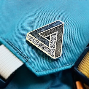 May include: A silver-coloured pin in the shape of a Penrose triangle, a geometric optical illusion. The pin features intricate etched patterns in black and grey, set against a teal fabric background. The pin is a unique accessory.