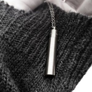 May include: A silver cylindrical pendant hangs from a silver chain. The pendant is suspended over a dark gray knitted fabric.