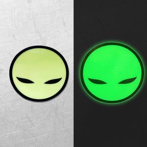 Op de afbeelding: Twee ronde stickers met een alien gezicht ontwerp. Eén sticker is lichtgroen met een zwarte omtrek en zwarte ogen, de andere gloeit felgroen met een zwarte omtrek en zwarte ogen. De achtergrond is verdeeld, met een lichtgrijze textuur aan de linkerkant en zwart aan de rechterkant.