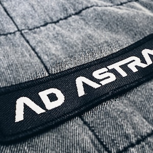 May include: A black patch with white stitching that reads "AD ASTRA" on a grey fabric background.
