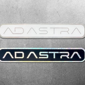以下が含まれることがあります： 長方形で角が丸い2つのステッカー。それぞれに「ADASTRA」の文字が表示されています。1つは銀色の文字が入った白、もう1つは虹色の文字が入った黒です。どちらも灰色の表面にあります。