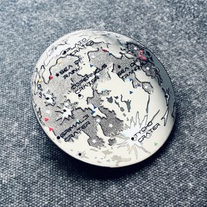 Op de afbeelding: Een zilverkleurige pin met een gedetailleerde kaart van de maan. De pin bevat de namen van verschillende kraters en zeeën, waaronder "Tycho Crater", "Grimaldi Crater", "Copernicus Crater" en "Sea of Rains".