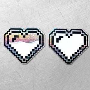 Könnte beinhalten: Zwei pixelige Herzaufkleber. Einer hat ein holografisches, irisierendes Finish mit rosa und weißen Farbtönen, der andere ist einfarbig weiß. Beide haben schwarze Umrisse und einen silbernen Glitzerrahmen. Die Aufkleber befinden sich auf einer strukturierten, grauen Oberfläche.