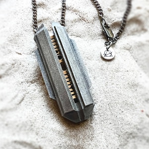 Puede incluir: Un collar con colgante plateado con un diseño geométrico. El colgante tiene forma rectangular con una estructura detallada y en capas y un interior dorado. La cadena del collar es de color plomo oscuro y está sobre arena.