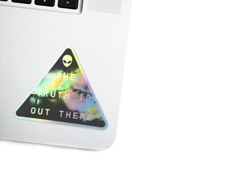 May include: A holographic sticker with a black triangle shape. The sticker has a pixelated design with a green alien face at the top and the text "THE TRUTH IS OUT THERE" in white.