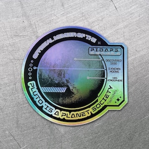 Könnte beinhalten: Ein holografischer Aufkleber mit kreisförmigem Design. Der Aufkleber zeigt eine Darstellung von Pluto mit dem Text "PLUTO IS A PLANET SOCIETY" und "OFFICIAL MEMBER OF THE P.I.D.A.P.S." Zusätzlicher Text lautet "DISCOVERED 1930", "5 KNOWN MOONS" und "HAS ATMOSPHERE."