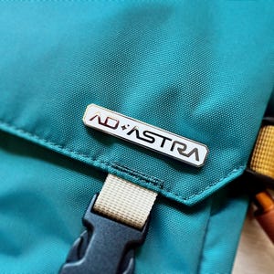 May include: Close-up of a teal backpack with a silver rectangular pin that reads "AD ASTRA" in black letters. The backpack has a black buckle and a tan strap. A yellow and orange strap is visible.