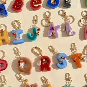Op de afbeelding: Een collectie kleurrijke, glanzende hars alfabet sleutelhangers, elk voorzien van een goudkleurige karabijnhaak en een kleine bedel zoals een aardbei, honingraat, schelp of zeester. De letters zijn uitgevoerd in diverse effen kleuren zoals rood, blauw en geel.