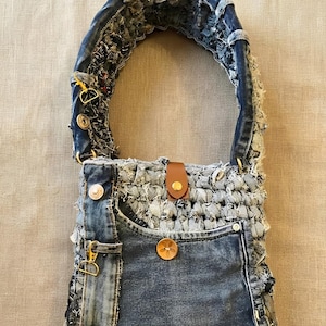 Pode incluir: Uma bolsa de ombro feita de tecido jeans azul reaproveitado com uma superfície desfiada e texturizada. A bolsa inclui uma alça grossa e arredondada de jeans, uma aba de fecho em couro marrom com um botão de pressão dourado.