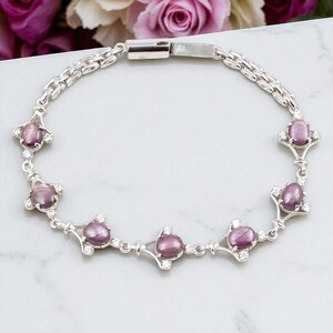 Könnte beinhalten: Ein silbernes Armband mit ovalen, purpurrosa Edelsteinen und kleinen, klaren Akzentsteinen. Das Armband hat ein filigranes Design mit einem Kettenglied-Armband und dekorativen Fassungen um jeden Edelstein. Das Armband wird auf einer weißen Marmoroberfläche präsentiert.
