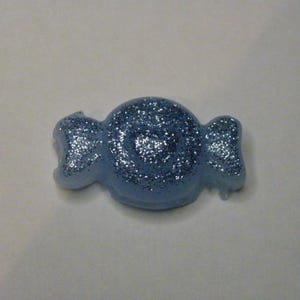 Può includere: Un ornamento a forma di caramella azzurra, ricoperto di glitter argentati. L'ornamento ha un design a caramella avvolta con estremità arrotondate e un corpo centrale circolare. I glitter aggiungono un effetto scintillante.