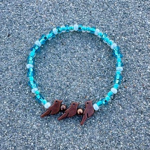 Puede incluir: Una pulsera de cuentas con cuentas turquesas y blancas. Tres dijes de pájaros marrones están centrados en la pulsera. La pulsera se muestra sobre una superficie gris texturizada.