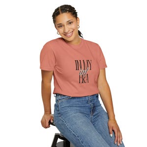 May include: A coral-colored t-shirt with the text "IN MY iof ERA" in black. The shirt is short-sleeved and worn with blue jeans. The person is sitting on a black stool.