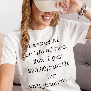 May include: White t-shirt with the text "I asked AI for life advice. Now I pay $20.00/month for enlightenment." A beige baseball cap is worn. The shirt is a casual, everyday wear item.