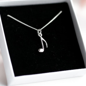 Può includere: Una collana d'argento con un ciondolo a forma di nota musicale, presentata in una scatola bianca con interno nero. La collana è delicata, con una catena sottile e un ciondolo a forma di croma stilizzata. Lo sfondo è sfocato, con toni rosa e bianchi.