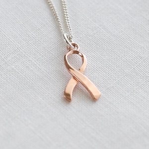 May include: A silver chain necklace with a rose gold ribbon pendant. The ribbon is a symbol of support and awareness. The necklace is shown on a white, textured background.