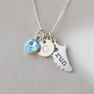 May include: Silver necklace with three charms: a round, light blue gemstone, a small disc with the letter "C", and a running shoe silhouette with the word "run". The necklace is on a silver chain.