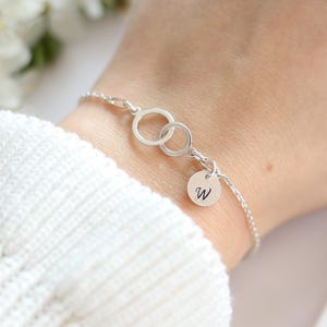 Op de afbeelding: Een zilveren armband met twee in elkaar grijpende cirkels en een kleine ronde bedel met de letter "W". De armband is delicaat en lijkt van sterling zilver te zijn. De achtergrond toont witte bloemen en een witte trui.