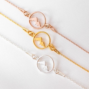 May include: Three necklaces with mountain pendant designs. One is silver, one is gold, and the other is rose gold. Each pendant features a mountain silhouette within a circular frame, connected to a fine chain.