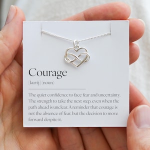 Puede incluir: Un collar plateado con un colgante en forma de corazón entrelazado con un símbolo de infinito, presentado en una tarjeta blanca. La tarjeta muestra la palabra "Coraje" y su definición.