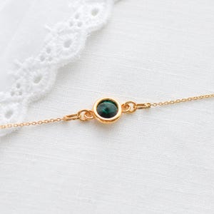 May include: A delicate gold necklace featuring a round, dark green gemstone pendant. The pendant is set in a gold bezel and connected to a fine gold chain. The necklace is displayed on a white, textured surface.
