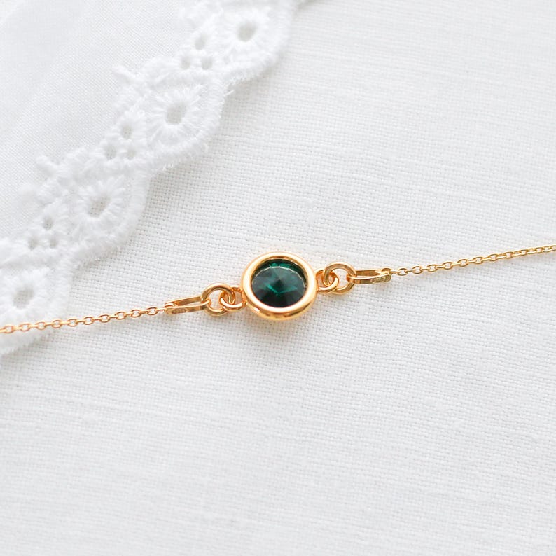 May include: A delicate gold necklace featuring a round, dark green gemstone pendant. The pendant is set in a gold bezel and connected to a fine gold chain. The necklace is displayed on a white, textured surface.
