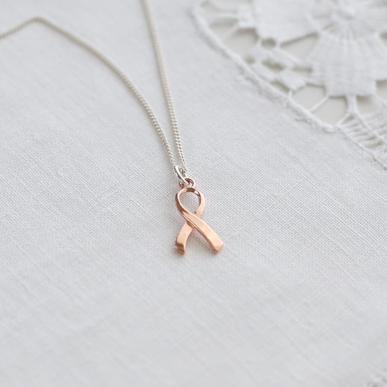 May include: A silver chain necklace with a rose gold ribbon pendant. The ribbon is a symbol of support and awareness. The necklace is shown on a white, textured background.