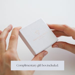 Op de afbeelding: Een witte geschenkdoos met een getextureerd oppervlak wordt door twee handen vastgehouden. De doos heeft een ros&eacute;gouden logo en de tekst "DIAMOND ROSE JEWELLERY". De tekst "Complimentary gift box included" wordt hieronder weergegeven.