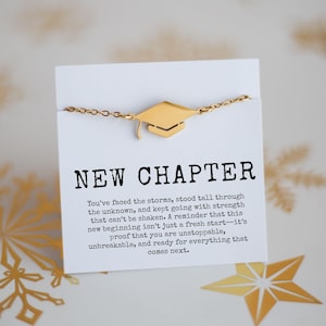 May include: A gold-colored graduation cap charm bracelet. The charm is a miniature mortarboard. The bracelet is on a white card with the words "NEW CHAPTER" and inspirational text. Gold star-shaped decorations are in the background.