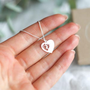 Op de afbeelding: Een zilveren hartvormige hangerketting met een uitgesneden ontwerp van babyvoetjes. De hartvormige bedel hangt aan een delicate ketting. De ketting wordt in een hand gehouden, met een vage achtergrond.