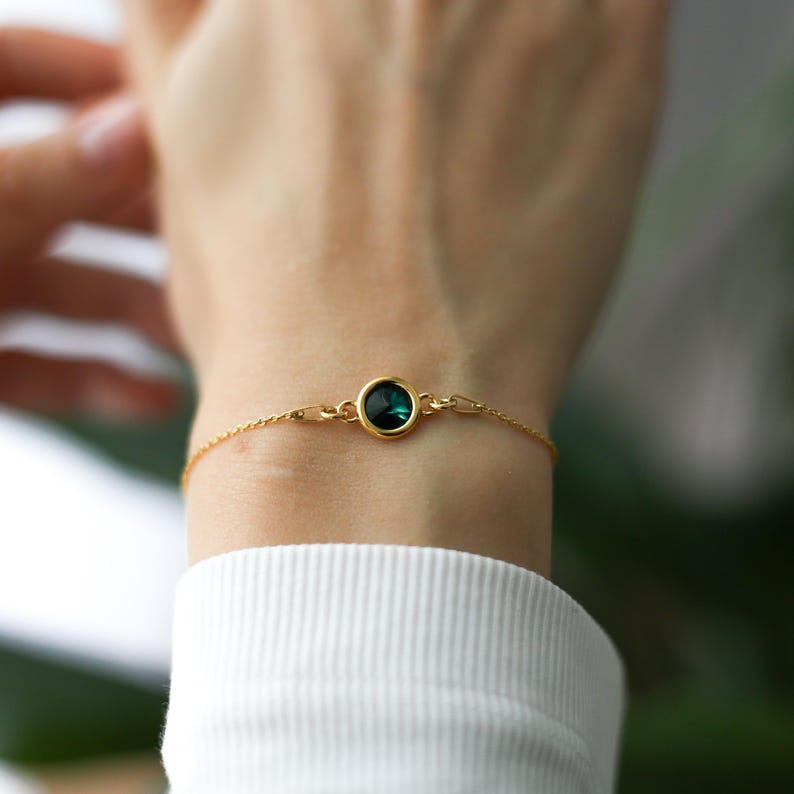 May include: A delicate gold bracelet featuring a round, dark teal gemstone. The bracelet is attached to a thin gold chain. The jewelry is worn on a wrist, with a white long-sleeved shirt in the background.