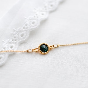 May include: A delicate gold necklace featuring a round, dark green gemstone pendant. The pendant is set in a gold bezel and is connected to a fine gold chain. The necklace is displayed on a white, textured surface.