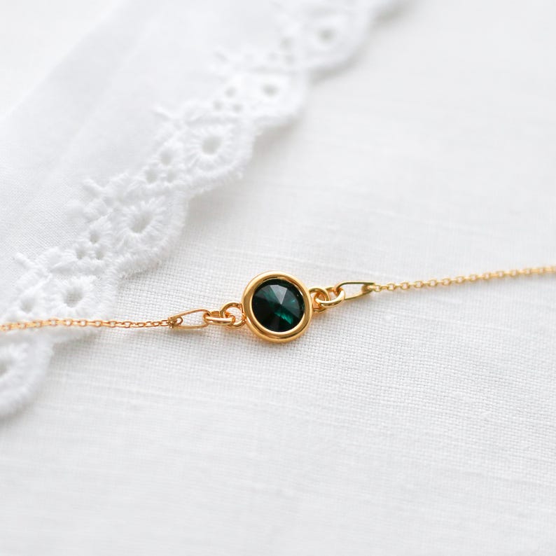 May include: A delicate gold necklace featuring a round, dark green gemstone pendant. The pendant is set in a gold bezel and is connected to a fine gold chain. The necklace is displayed on a white, textured surface.